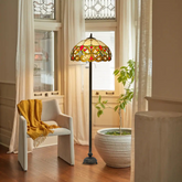 A Tiffany style 16 inch floor lamp with a multicolored stained glass shade, placed in a room with a chair and a plant, providing a warm glow.