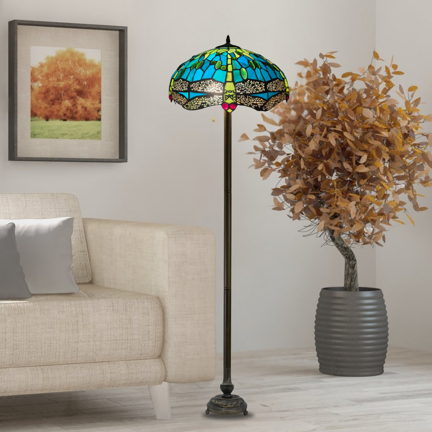 A Tiffany floor lamp with a green stained glass shade, placed in a room next to a sofa with a picture frame on the wall and a potted plant.