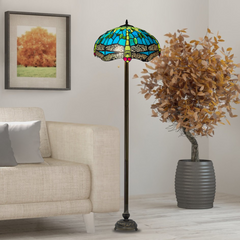 A Tiffany floor lamp with a green stained glass shade, placed in a room next to a sofa with a picture frame on the wall and a potted plant.