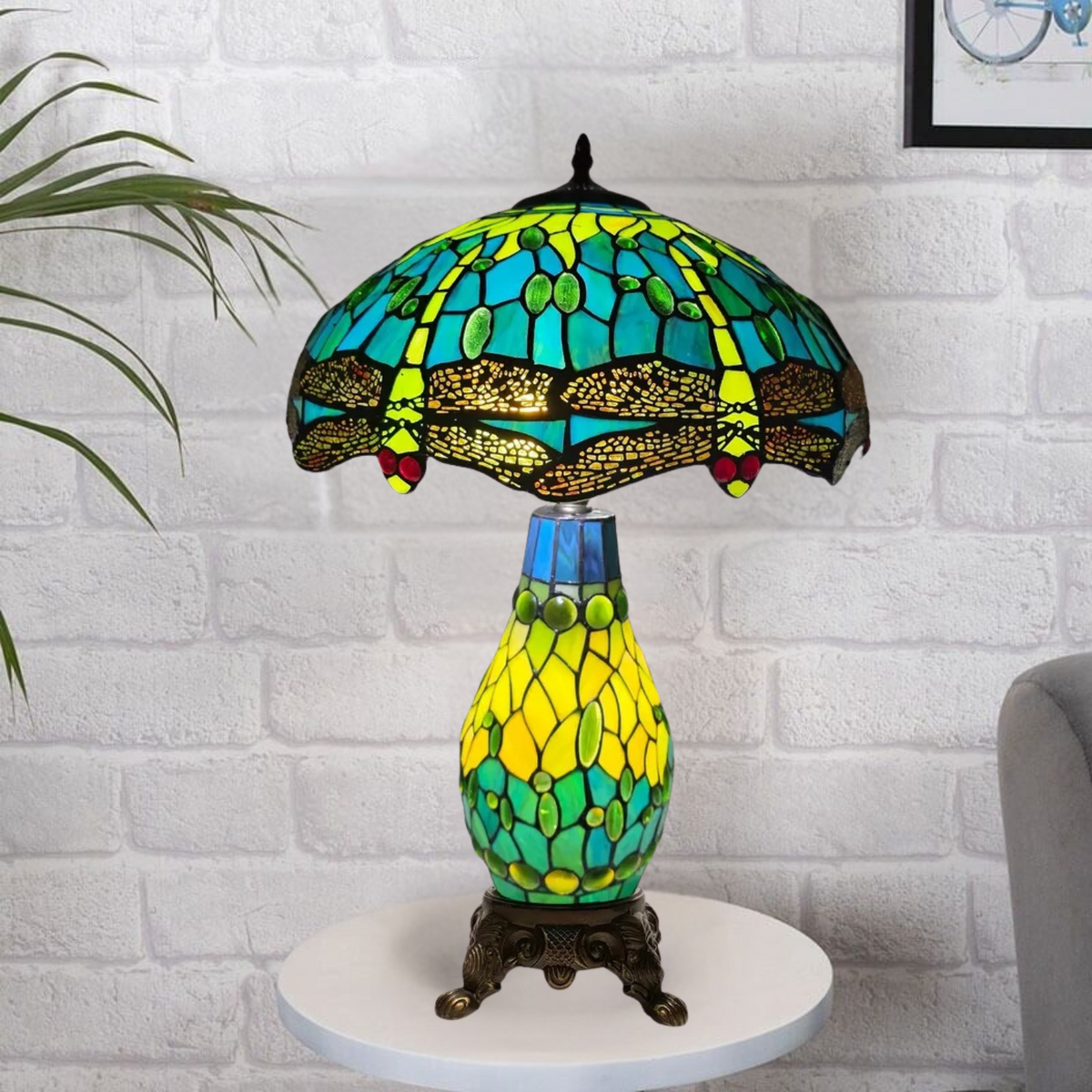 A colorful Tiffany style clustered table lamp with a stained glass shade and a metal base, placed on a white surface against a brick wall background.