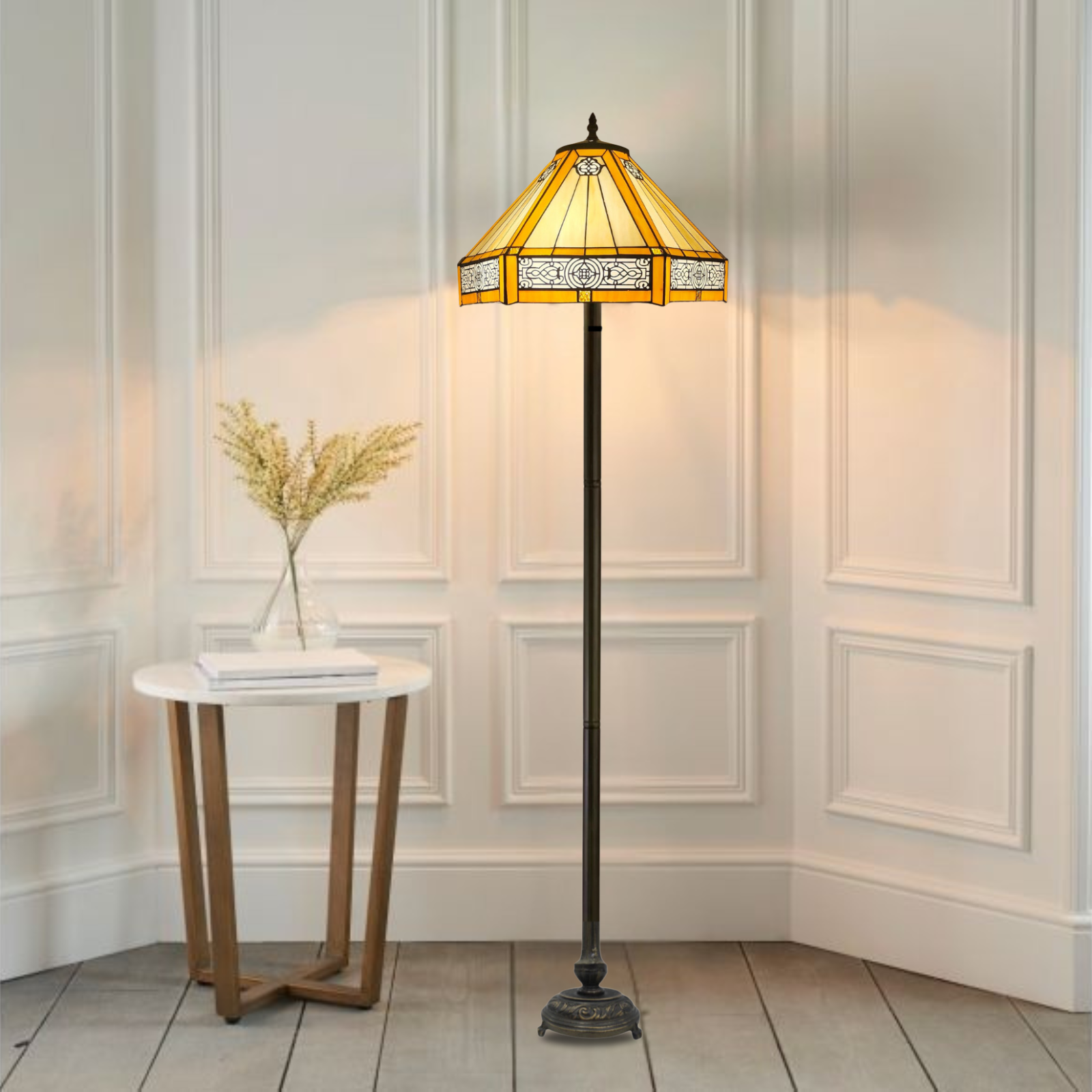 A yellow hexagonal Tiffany floor lamp with a handcrafted stained glass shade and a metal base, placed in a room next to a round white table with a vase of dried flowers on it.